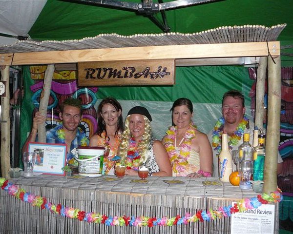 Guests in pirate-themed costumes behind a tiki bar labeled "RUMBar" at a Caribbean murder mystery party.