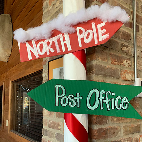 Holiday-themed North Pole and Post Office signs with snow-like decor on a candy cane-striped pole.