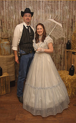 Couple dressed in Old West attire posing in a murder mystery setting with rustic props like hay bales and barrels.