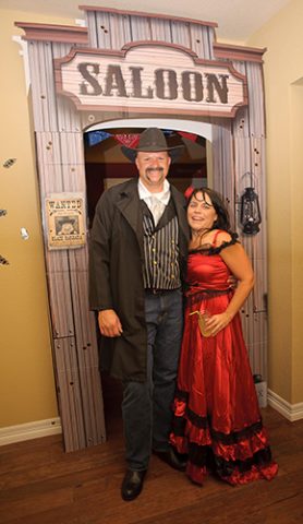 Man and woman in Old West costumes posing at a saloon-themed entrance with a 'WANTED' poster on the wall.