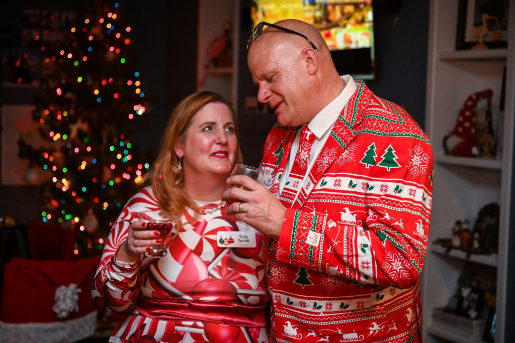 Couple in festive Christmas outfits holding drinks at a holiday party with a decorated tree in the background.