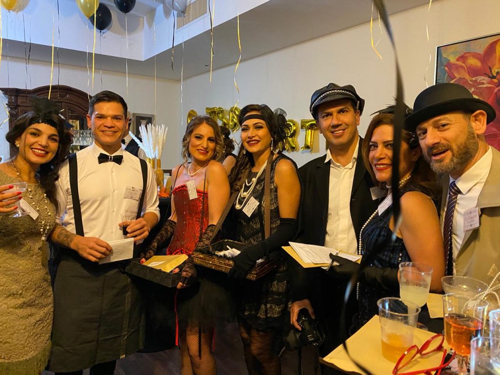 Group of people dressed in 1920s-themed attire, including flapper dresses, suspenders, pearls, and hats.