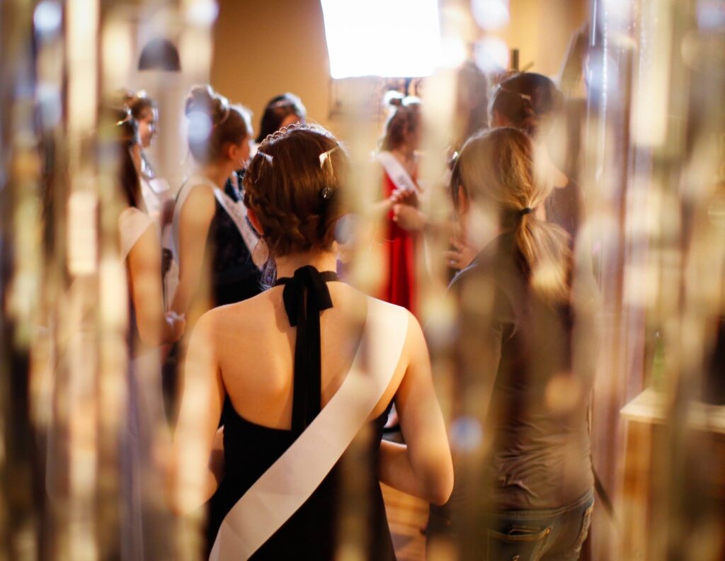 Night of Mystery | Sweet-16 Themes: From Glam To Mystery She’ll Never Forget A group of girls wearing sashes gathered together at a pageant-themed event, viewed through a shimmering tinsel curtain.