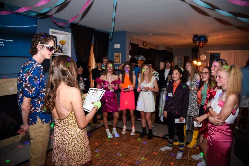 Night of Mystery | Sweet-16 Themes: From Glam To Mystery She’ll Never Forget A group of teens in retro 1980s attire gathered on a dance floor during a themed murder mystery party.