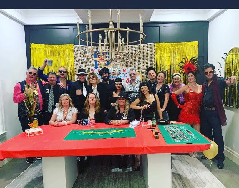 Night of Mystery | 30th Birthday Party Ideas To Throw The Best Mystery Of The Decade Women in Wild West costumes holding up party awards alongside a large group of guests gathered around a casino-themed table for a murder mystery event.
